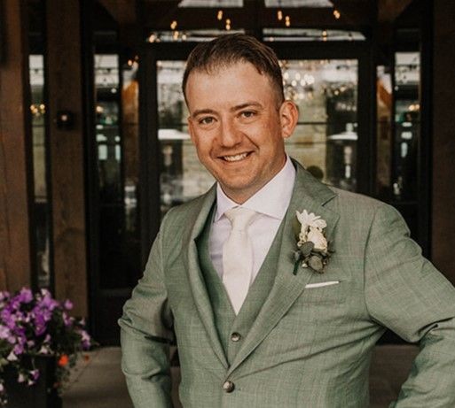 Man in green suit smiling, standing outdoors with flowers in the background.